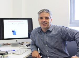 A portrait of Professor James Walters who is a middle-aged white man with short grey hair, he sits at a desk with a computer, smiling at the camera, and he is wearing a grey blue shirt.