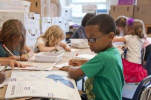 little black boy writing in a workbook in a busy classroom