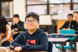 young asian boy smiling while sat at the front of a class