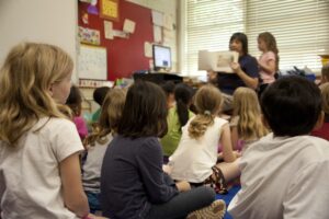 children sit on a classroom floor listening to a teacher reading from a book