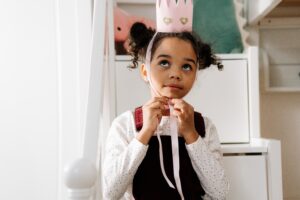 a little girl ties ribbon under her chin of a pink paper crown