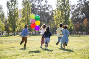 a group of children run through a field holding bright coloured balloons