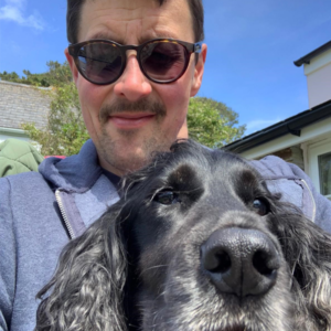 a photo of NCMH admin officer Mark, wearing sunglasses and a blue hoody and his black dog is in front of him close to the camera