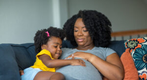 a pregnant black woman with her little girl sitting on the sofa touching her pregnant belly