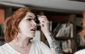 young woman with ginger hair and tattoos applying mascara in a mirror
