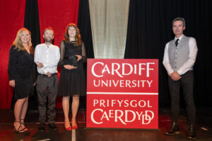 Two men and two women standing on stage with a large red square Cardiff University logo