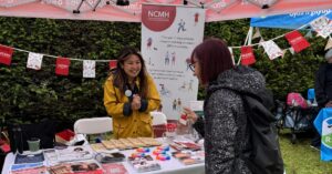 NCMH holding a stand at an event in Insole Court as part of the Every Woman Festival. A young woman with red hair speaks to a member of staff, Jess Yang, who has long brown hair and wears a yellow raincoat.