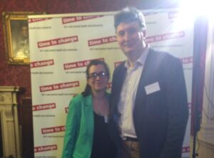 Professor Ian Jones and Laura, an NCMH Research Champion, stand together in front of a 'Time to Change' banner. Ian is tall with dark hair, a dark jacket and white shirt. Laura is shorter with dark hair and glasses wearing a bright green jacket. Sunlight is streaming into the top right corner of the image.