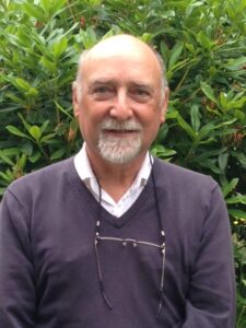 Peter is an older man with short grey hair and beard. He wears a purple jumper and a white shirt with glasses hanging around his neck.