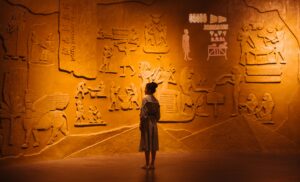 A young woman with dark hair stands in front of a wall lit in yellow. The wall has hieroglyphs on it.